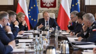 Premier i ministrowie podczas pilnego posiedzenia przy długim stole, w tle polskie i unijne flagi