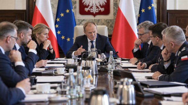 Premier i ministrowie podczas pilnego posiedzenia przy długim stole, w tle polskie i unijne flagi