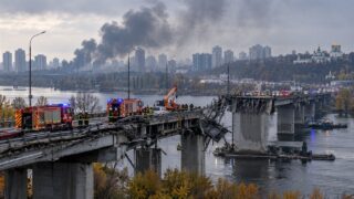 Uszkodzony most nad szeroką rzeką, służby ratunkowe i dym nad miastem
